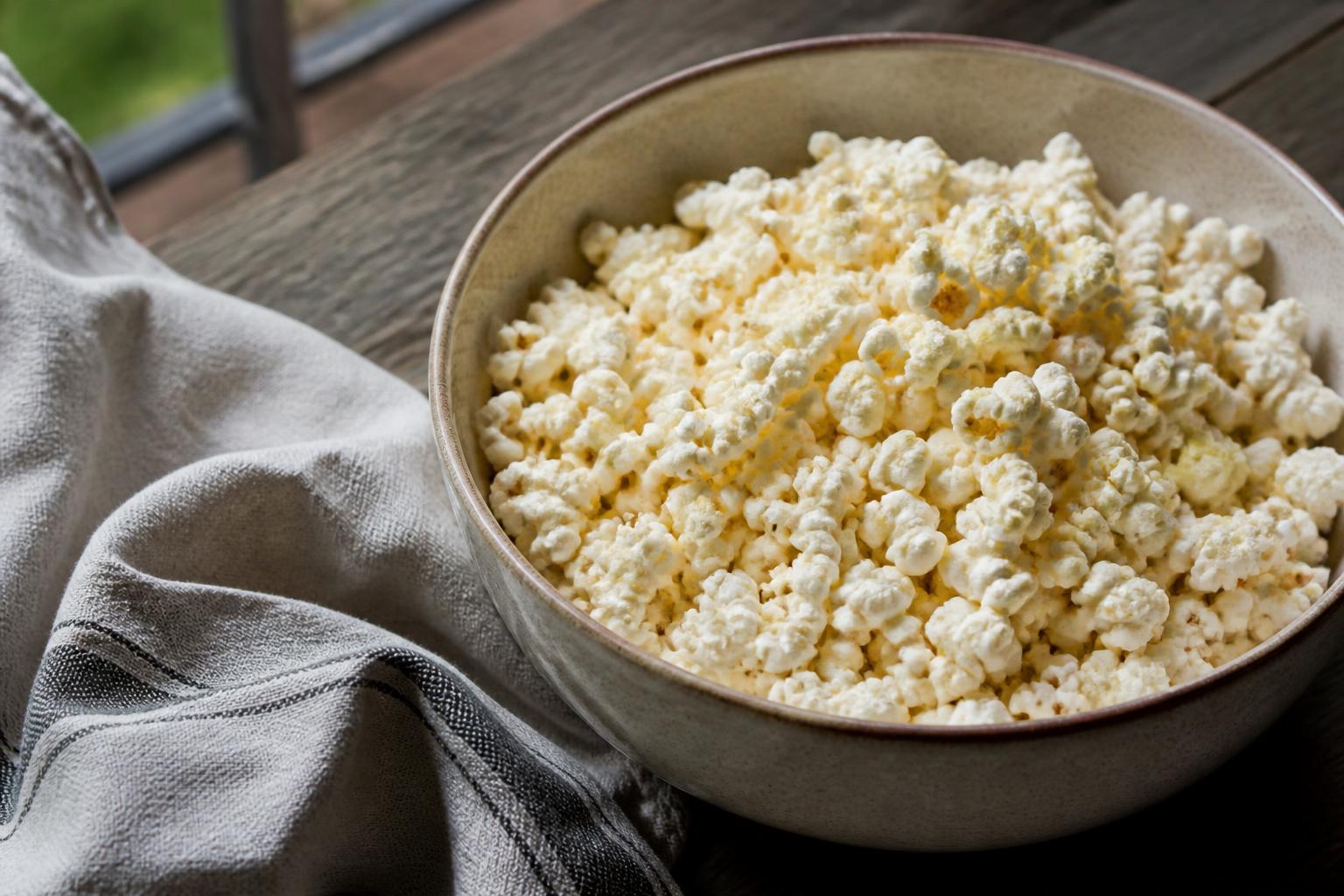 A bowl of fluffy, white popcorn sits on a wooden table against a soft-focus background