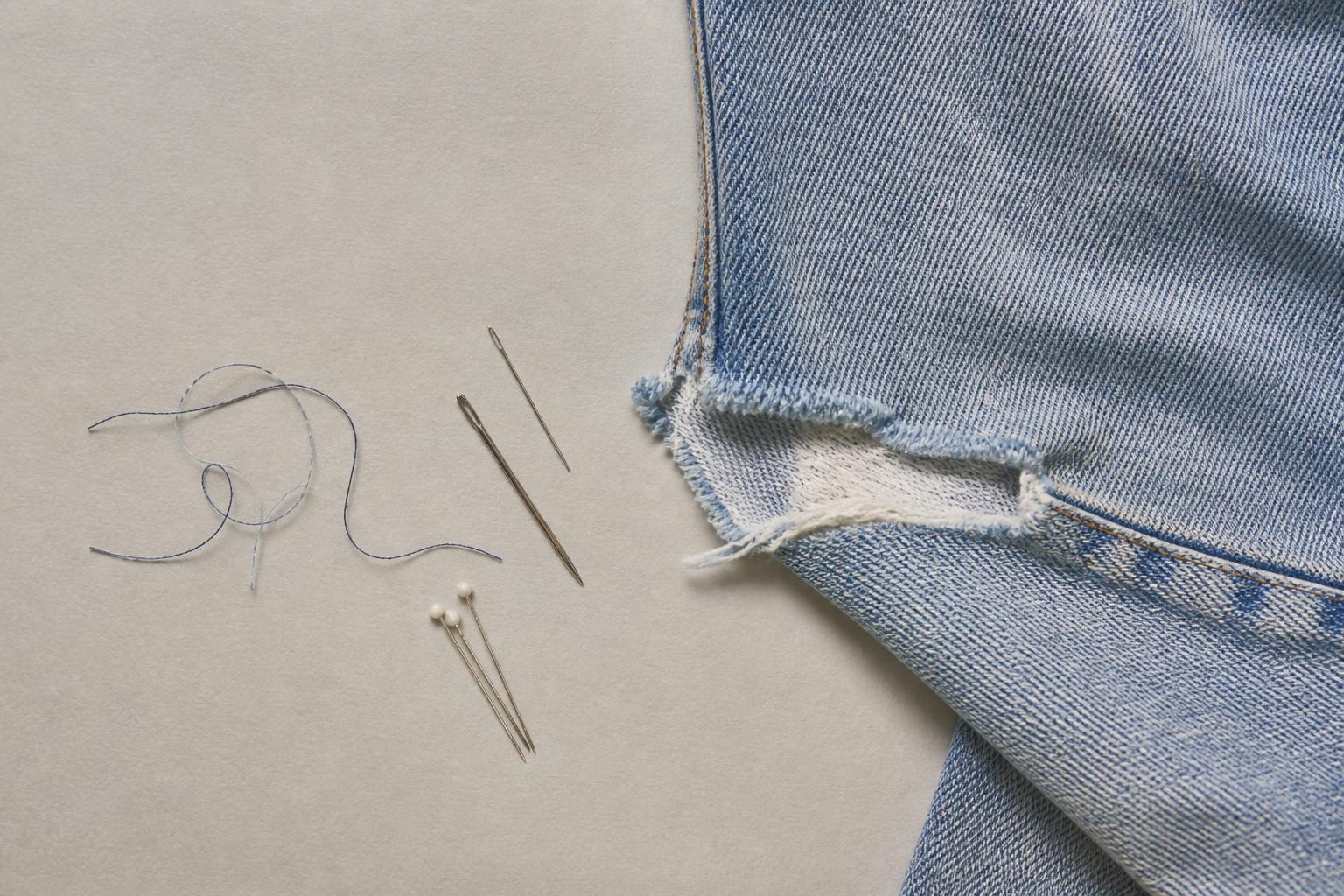 A tailor inspects a damaged denim jacket on a workbench cluttered with sewing tools and fabric swatches
