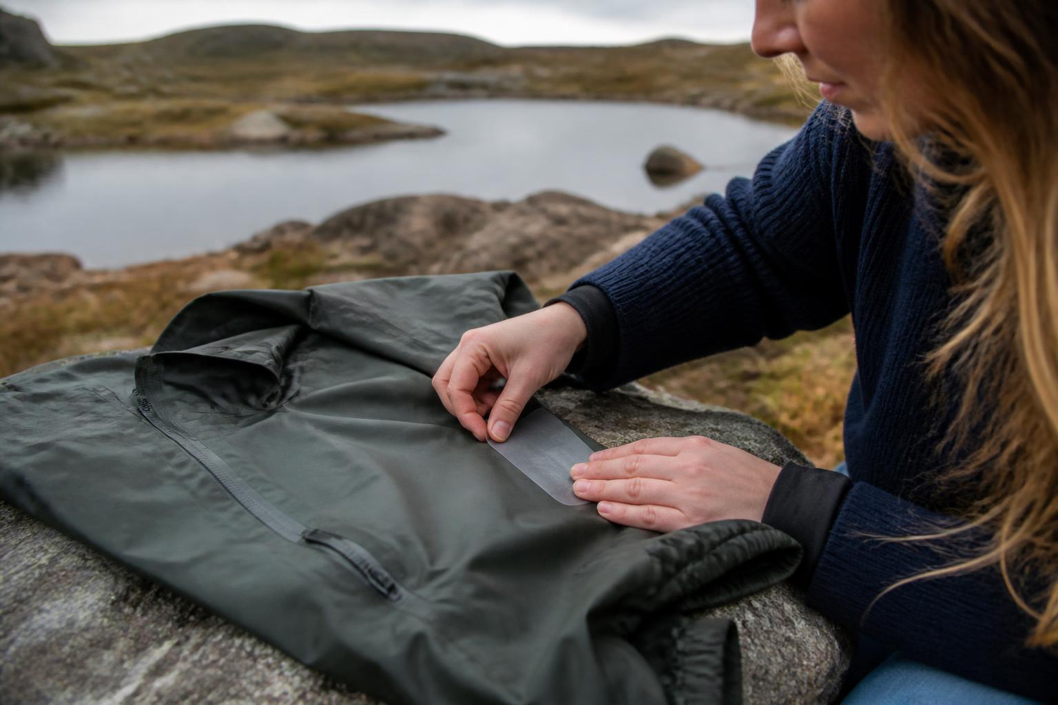 Close-up of a person applying a transparent adhesive patch to the frayed seam of a blue waterproof jacket