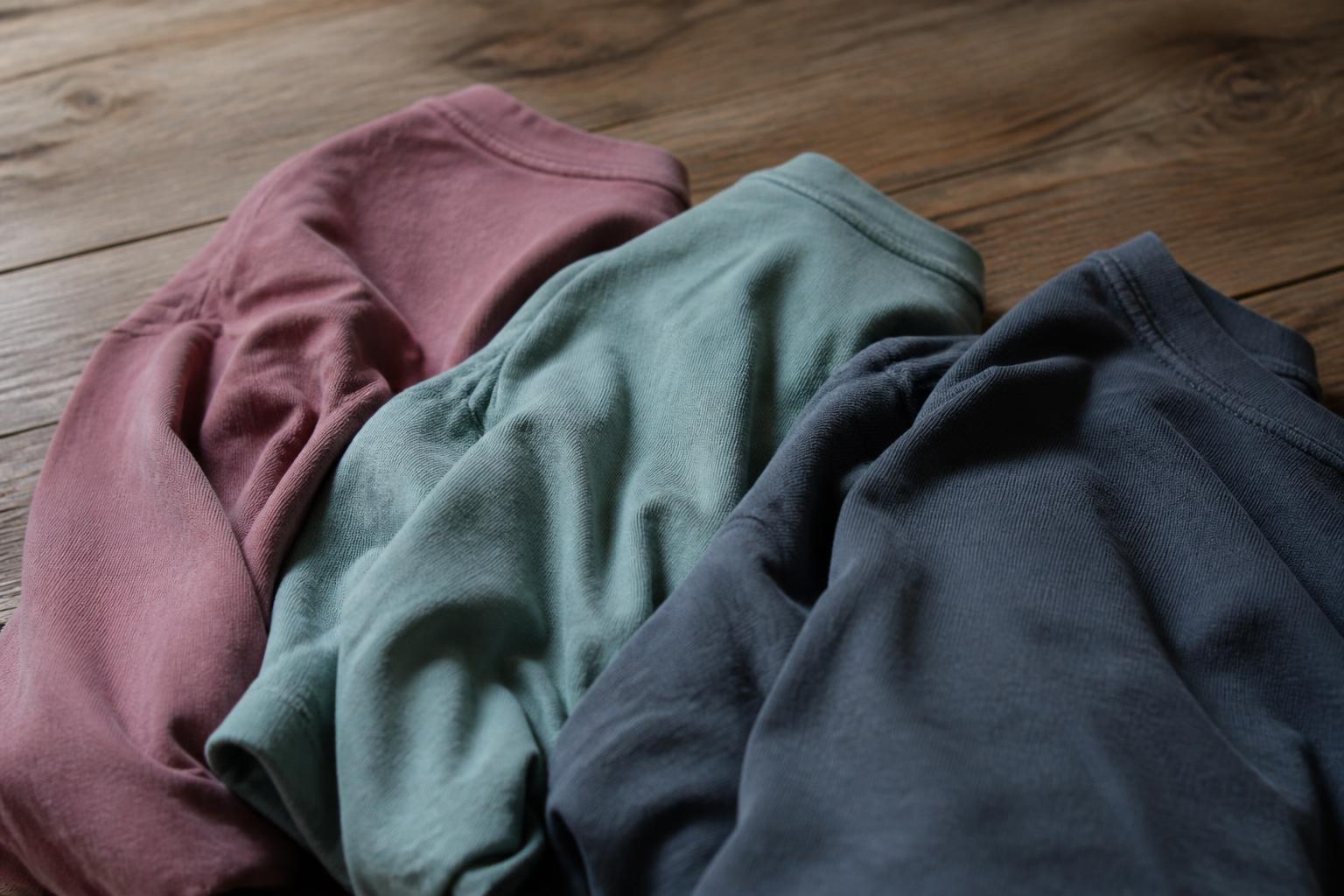 A stack of folded cotton t-shirts in various muted, unevenly dyed pastel shades resting on a wooden surface
